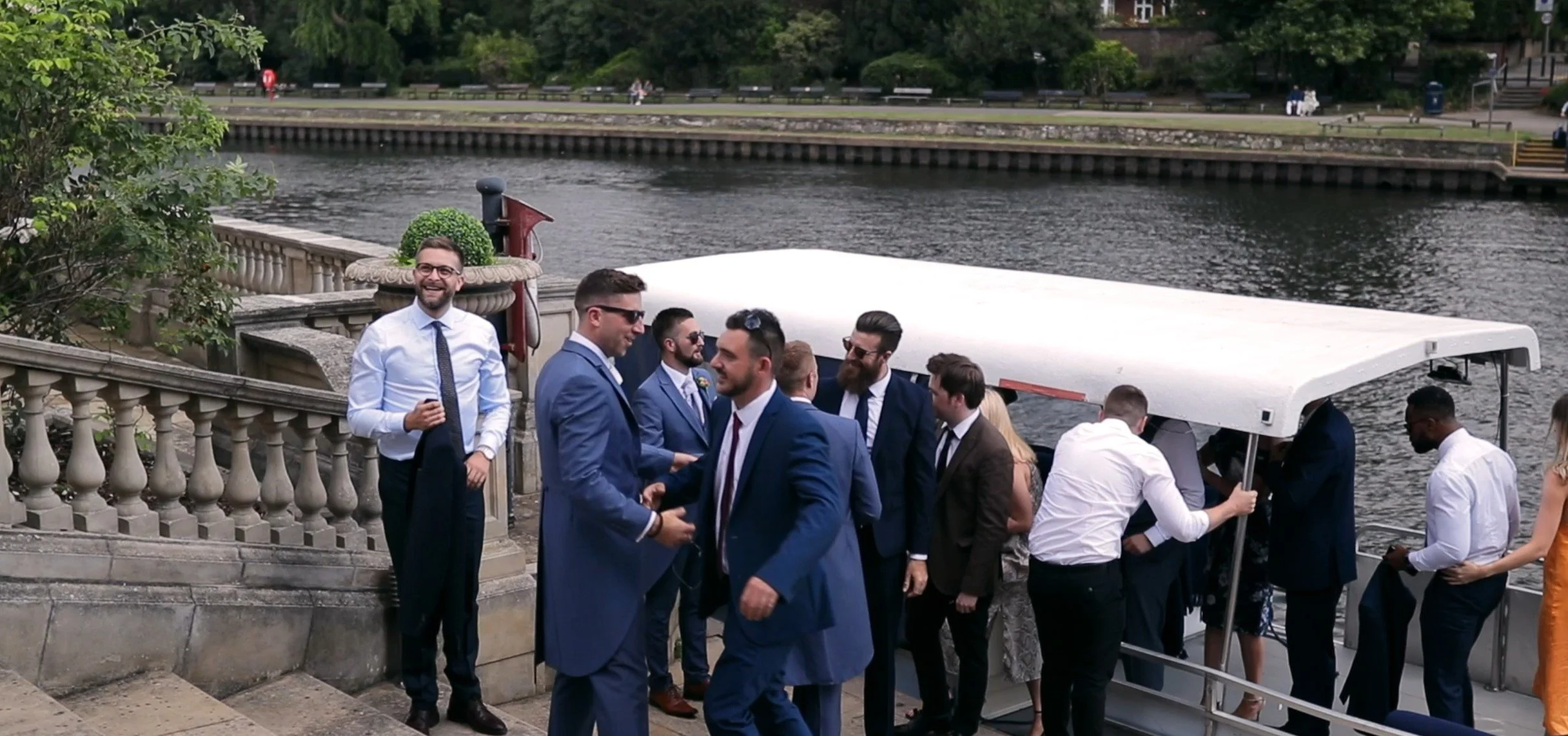Group of men in formal suits standing near river, some talking, some adjusting jackets, with a small boat docked nearby, and a park with trees and benches in the background.