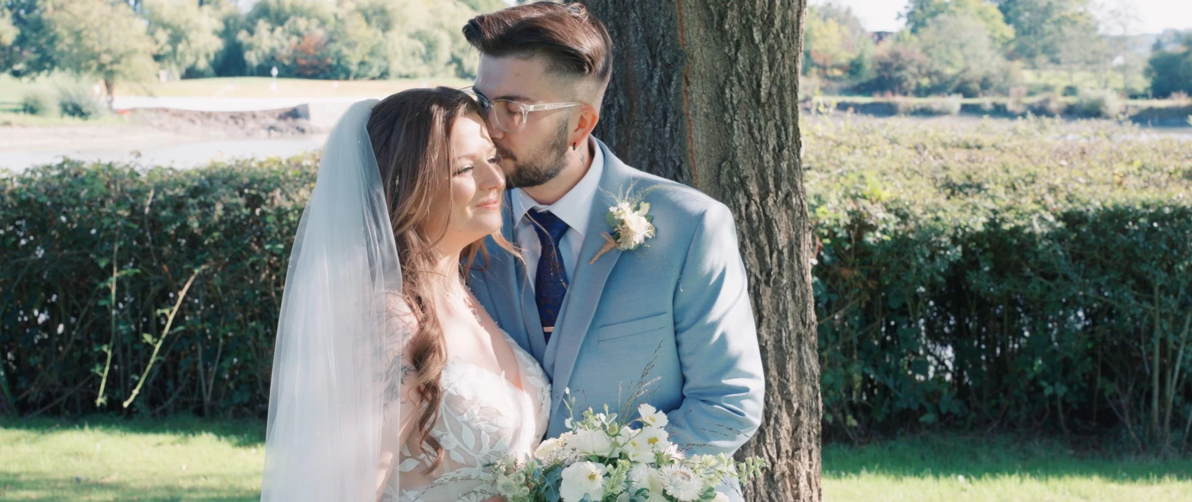 A bride and groom sharing a kiss in an outdoor setting during their wedding, with the groom wearing glasses and a light blue suit, and the bride holding a bouquet of white flowers.