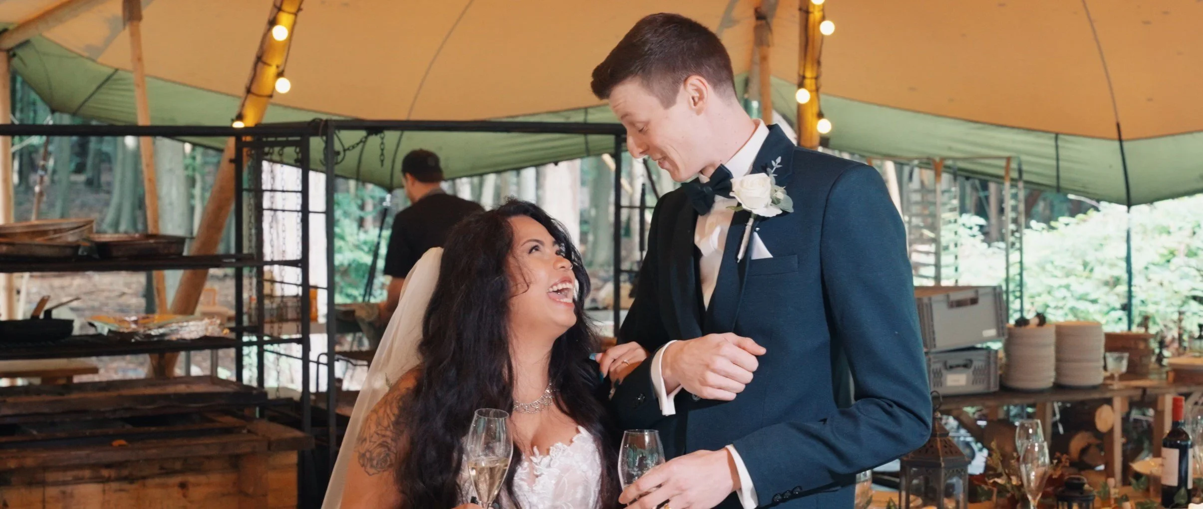 Bride and groom sharing a joyful moment, with the bride sitting and the groom standing inside a tent at a wedding reception.