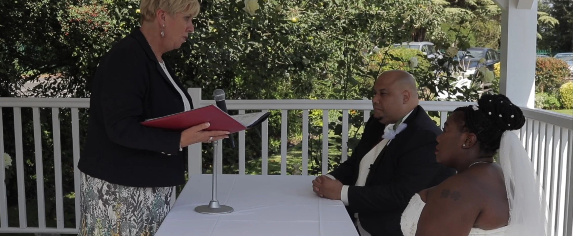 A woman officiant standing and reading vows to a seated couple during a wedding ceremony on a porch with greenery in the background.