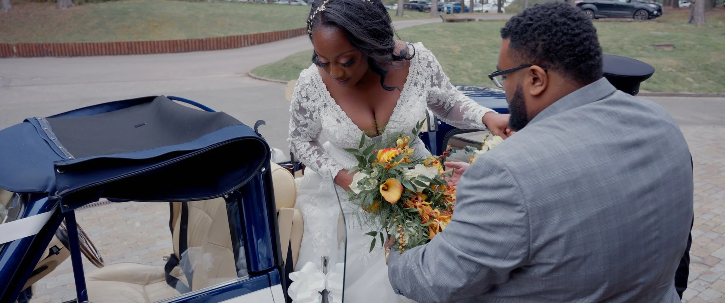 A bride in a white lace wedding dress holding a bouquet of flowers, leaning into a man in a gray suit who is giving her a flower from his hand beside a blue vintage car.