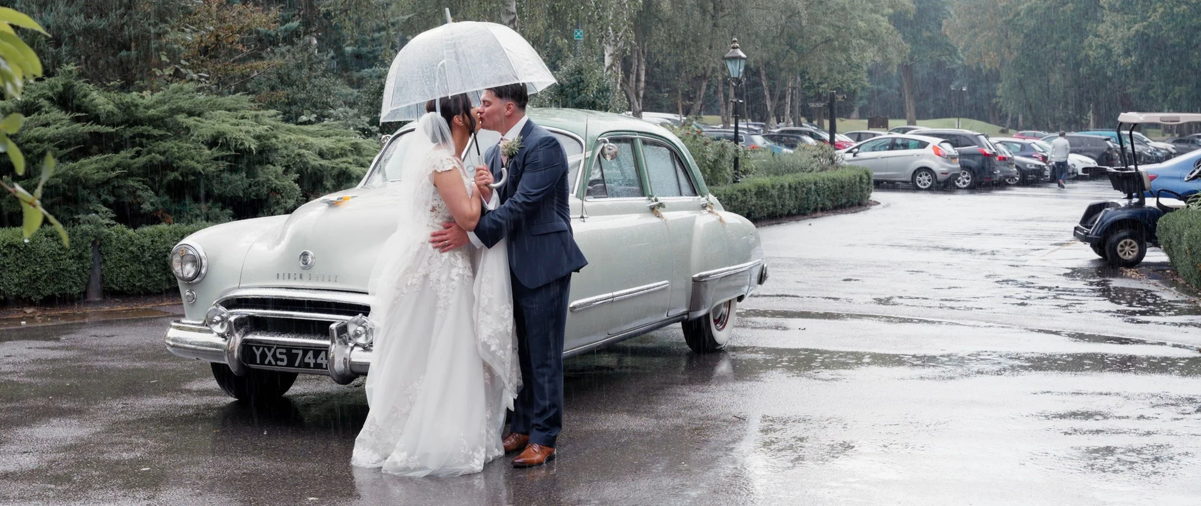 A bride and groom sharing a kiss under an umbrella on a rainy day, standing next to a vintage white car in a parking lot, with numerous cars and greenery in the background.