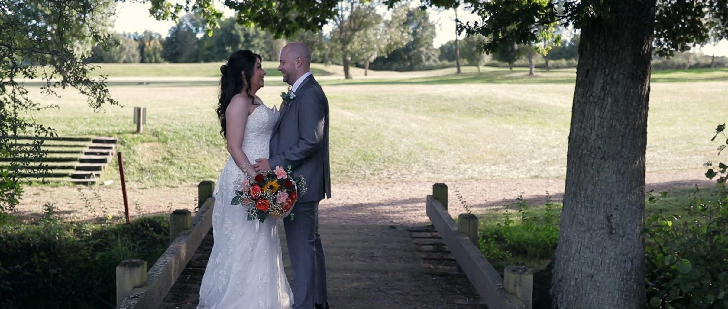 Bride and groom standing face to face on a small wooden bridge in a park, holding hands, with trees and open grass in the background.