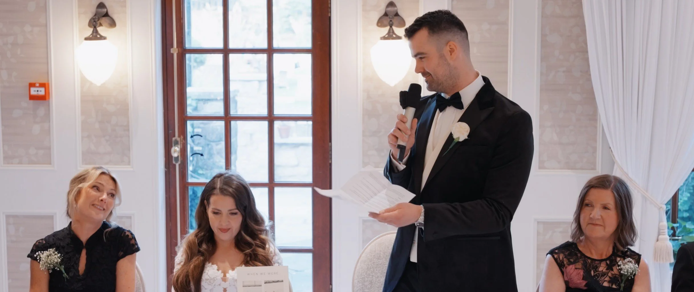A man in a black tuxedo giving a speech at a wedding reception, holding a microphone and reading from a paper, surrounded by seated women dressed in formal attire.