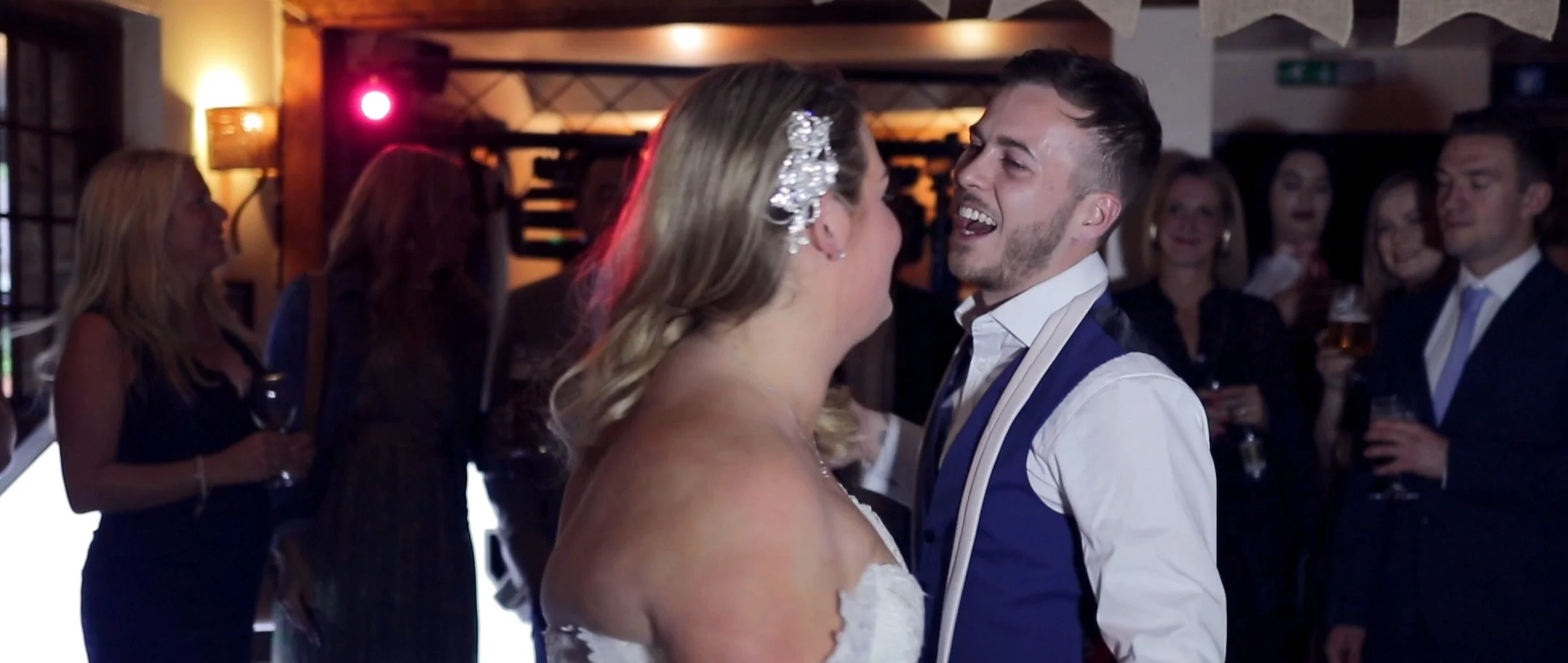 A bride and groom dancing and smiling at their wedding reception, with guests in the background holding drinks and enjoying the celebration.