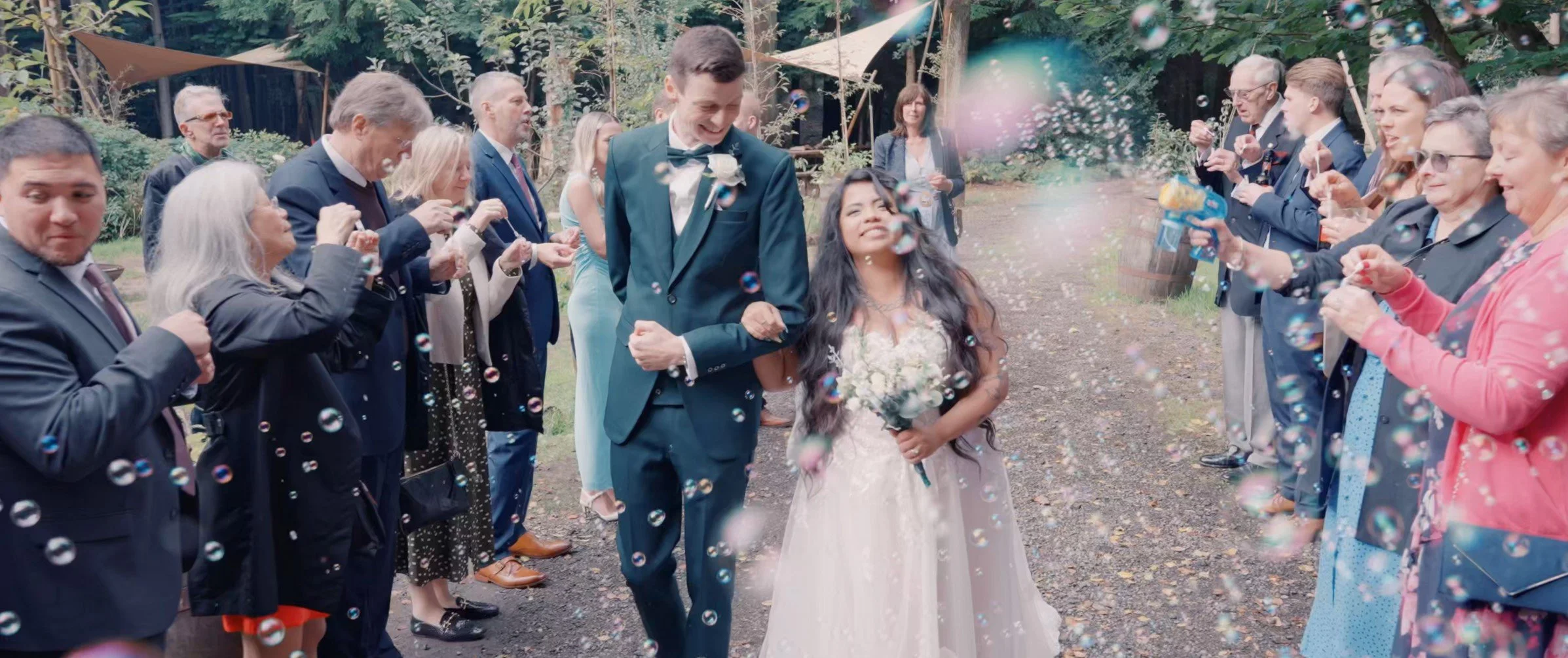A bride and groom walk down the outdoor aisle surrounded by family and friends, with bubbles floating around during their wedding celebration.