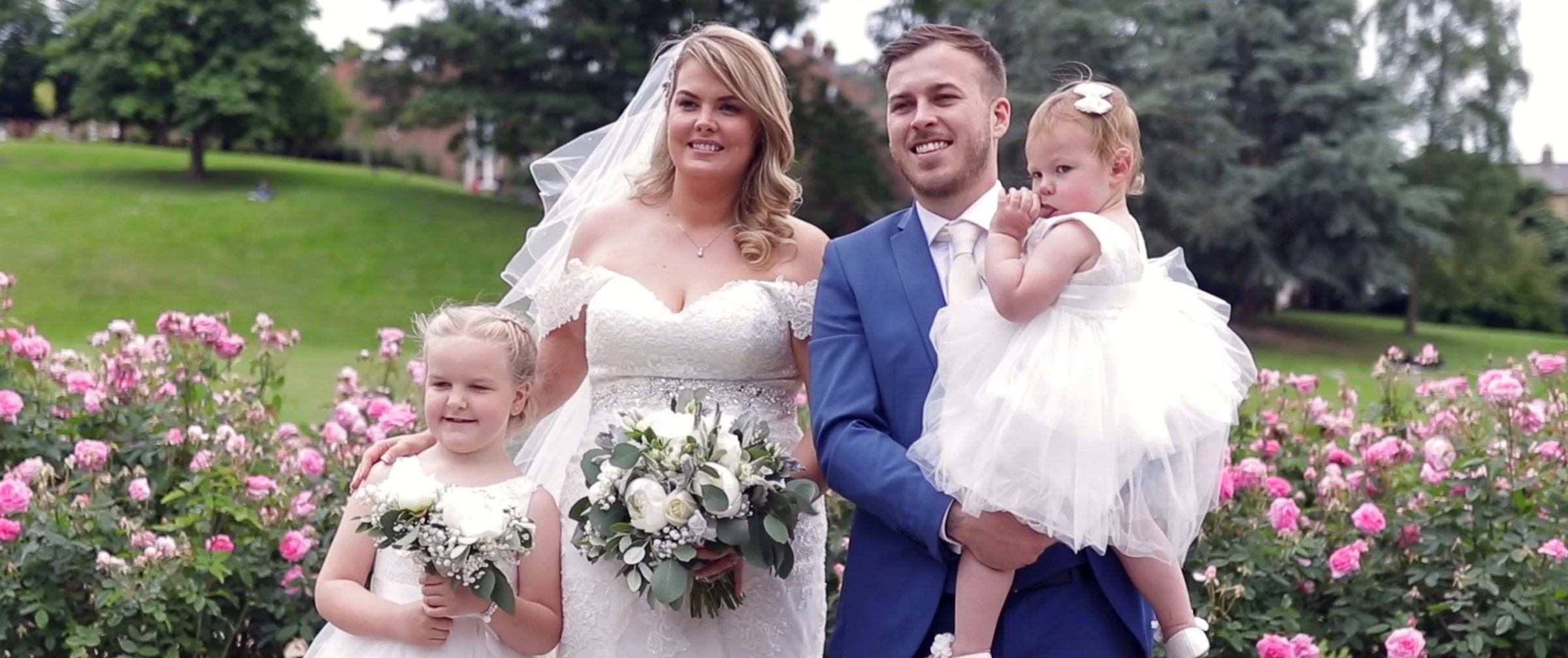 A bride in a lace wedding dress holding a bouquet and smiling, standing next to a groom in a blue suit holding a young girl in a white dress, with another girl in a white dress holding a bouquet in front, in a garden with pink flowers and trees.