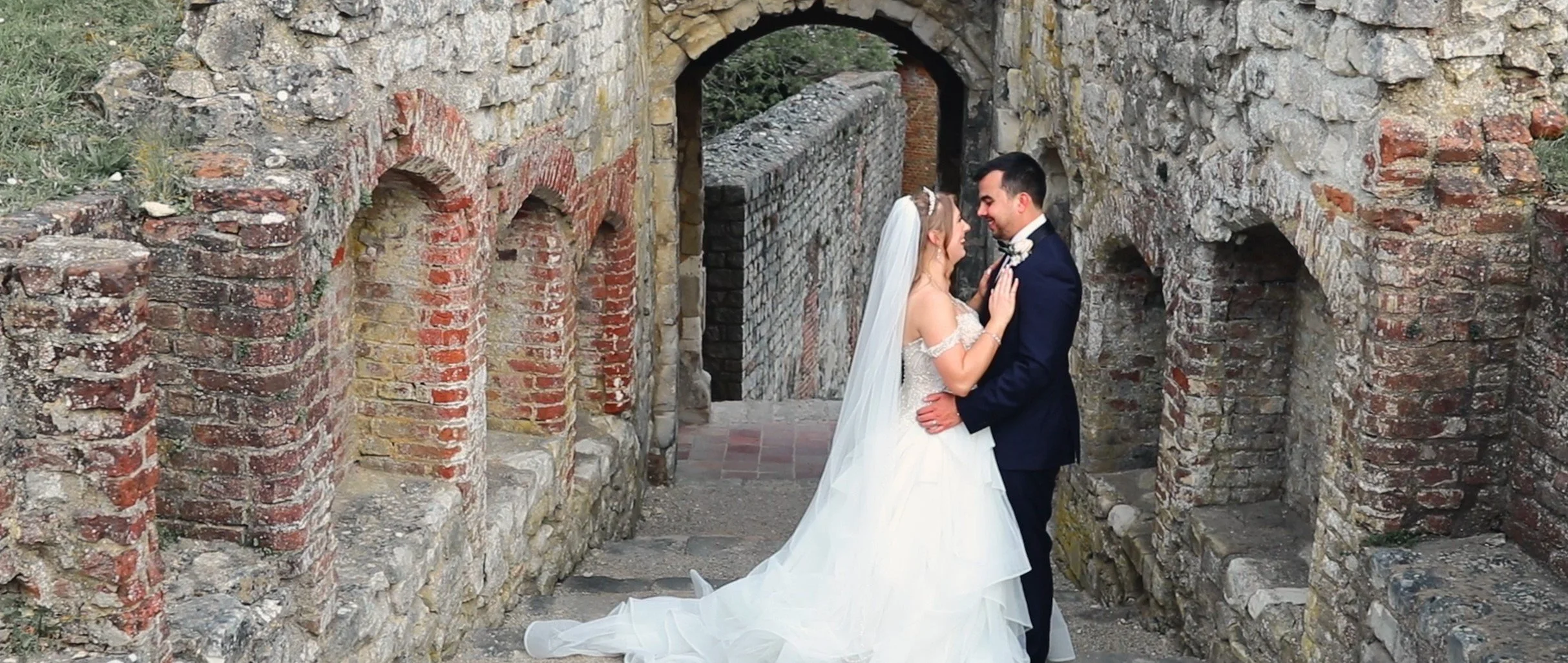 A bride and groom in wedding attire standing close together, smiling, in front of a rustic stone and brick arched structure.