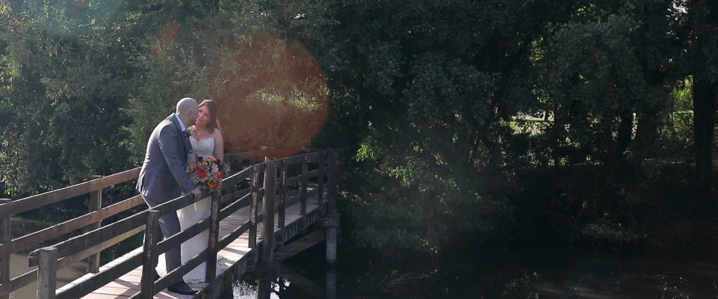 A bride and groom standing on a wooden bridge surrounded by trees, sharing a romantic moment with the groom holding a bouquet of flowers.