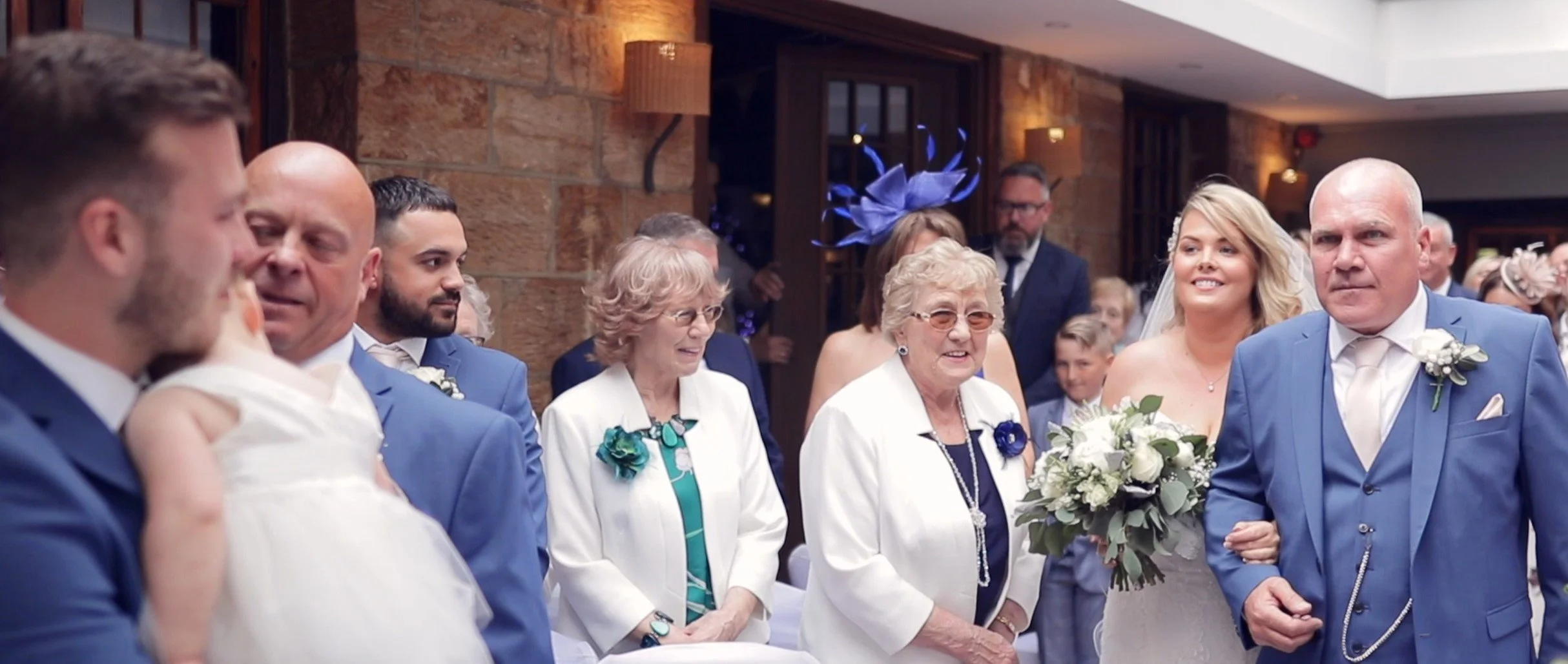 A wedding reception with guests, including elderly women, dressed in formal attire, some smiling, with a bride holding a bouquet, in a decorated indoor venue with brick walls and warm lighting.