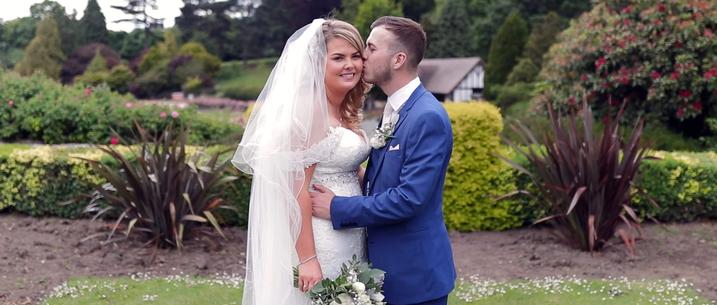 A bride and groom on their wedding day in a garden, with the groom kissing the bride on the cheek. The bride is wearing a wedding dress with a veil, holding a bouquet, and the groom is in a blue suit. There are green bushes and colorful flowers in th