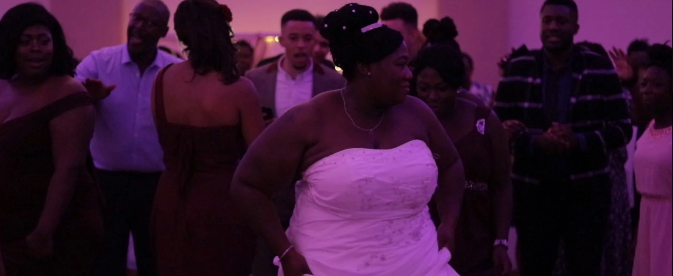 Group of people dancing and socializing at an indoor event with purple lighting, including women in dresses and men in shirts and jackets.