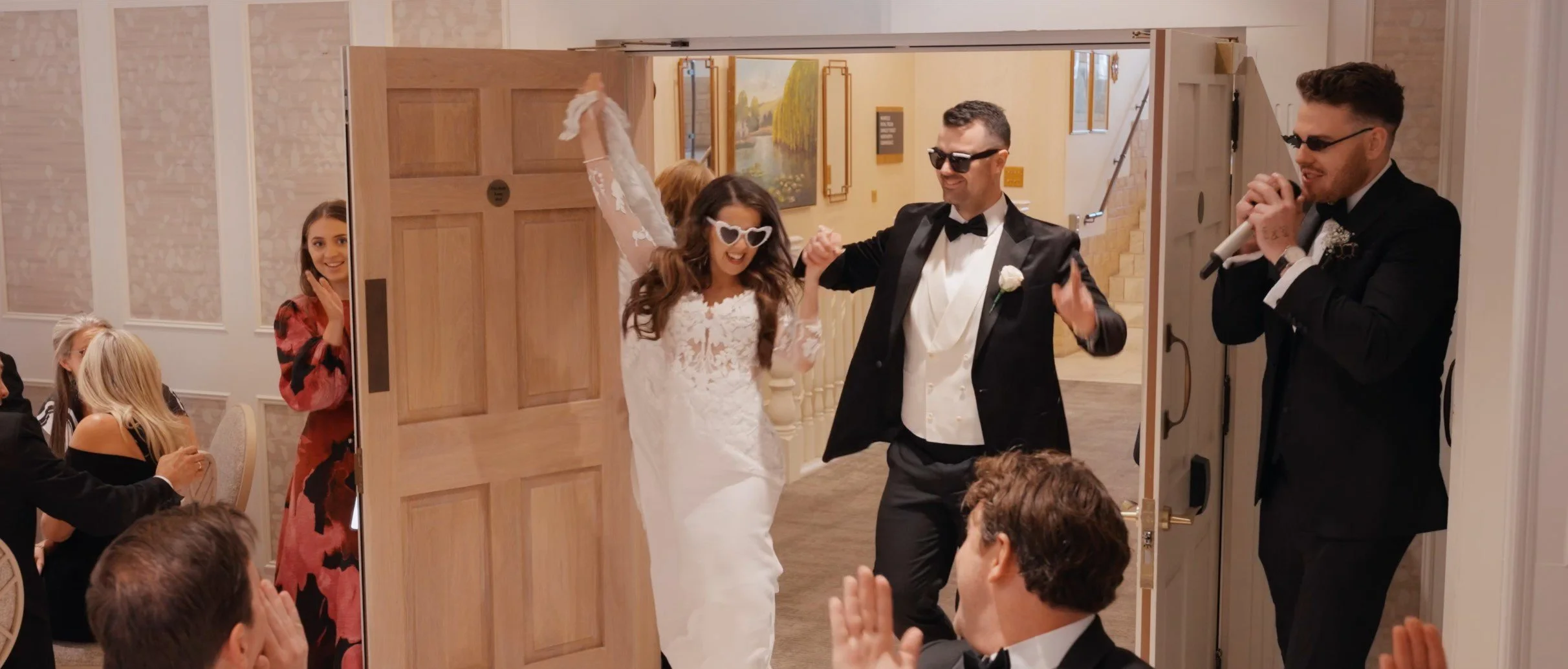 A bride and groom enter a wedding reception, holding hands and smiling. The bride wears a white lace wedding dress and fun heart-shaped sunglasses. The groom wears a black tuxedo with a bow tie and sunglasses. Guests are seated around, clapping and s