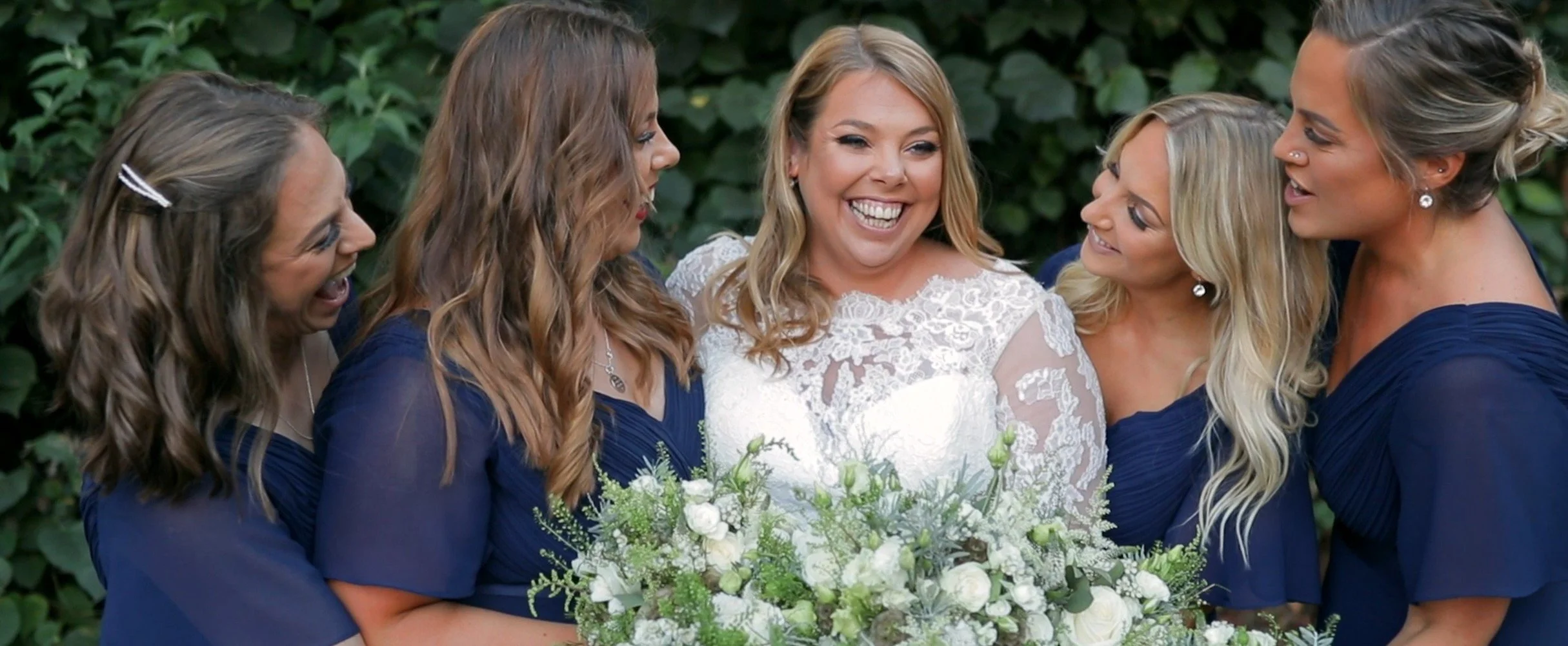 A bride in a white lace wedding dress holding a bouquet, surrounded by five bridesmaids in navy blue dresses, all smiling and looking at each other outdoors with greenery in the background.