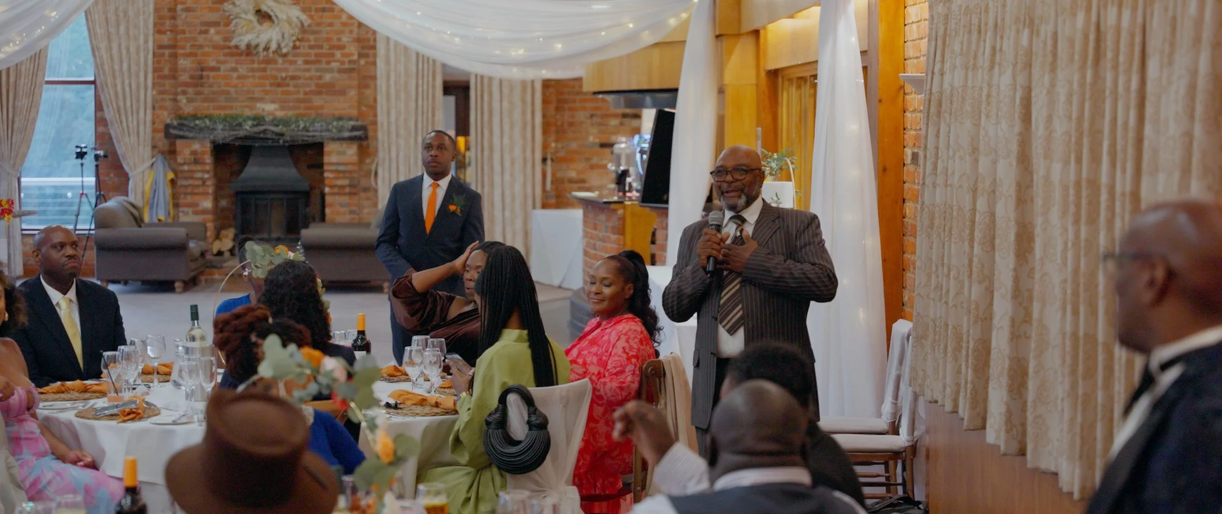 A man in a striped suit holding a microphone and speaking at a formal gathering, with seated guests listening, in a decorated indoor venue with brick walls and curtains.