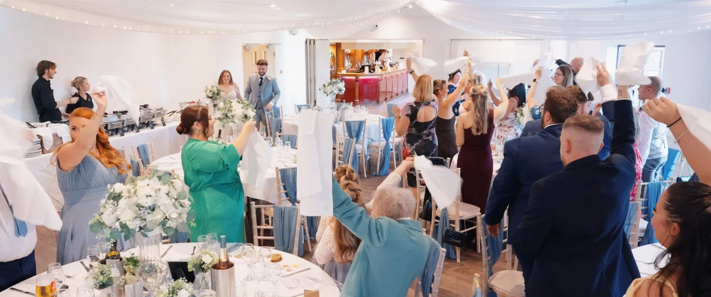 People celebrating at a wedding reception, standing and raising napkins, with the bride and groom in the background at the head table.