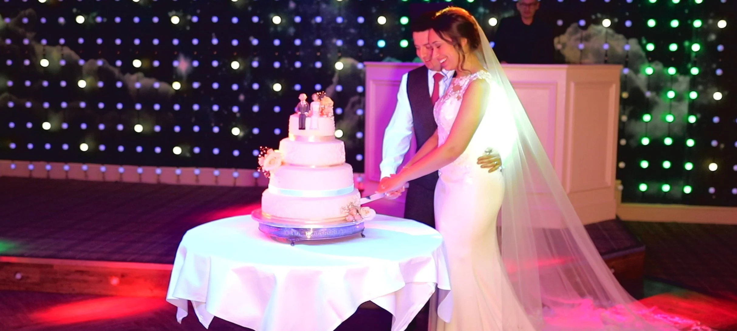 A bride and groom cutting their wedding cake together during their wedding reception, with festive lighting and decorations in the background.