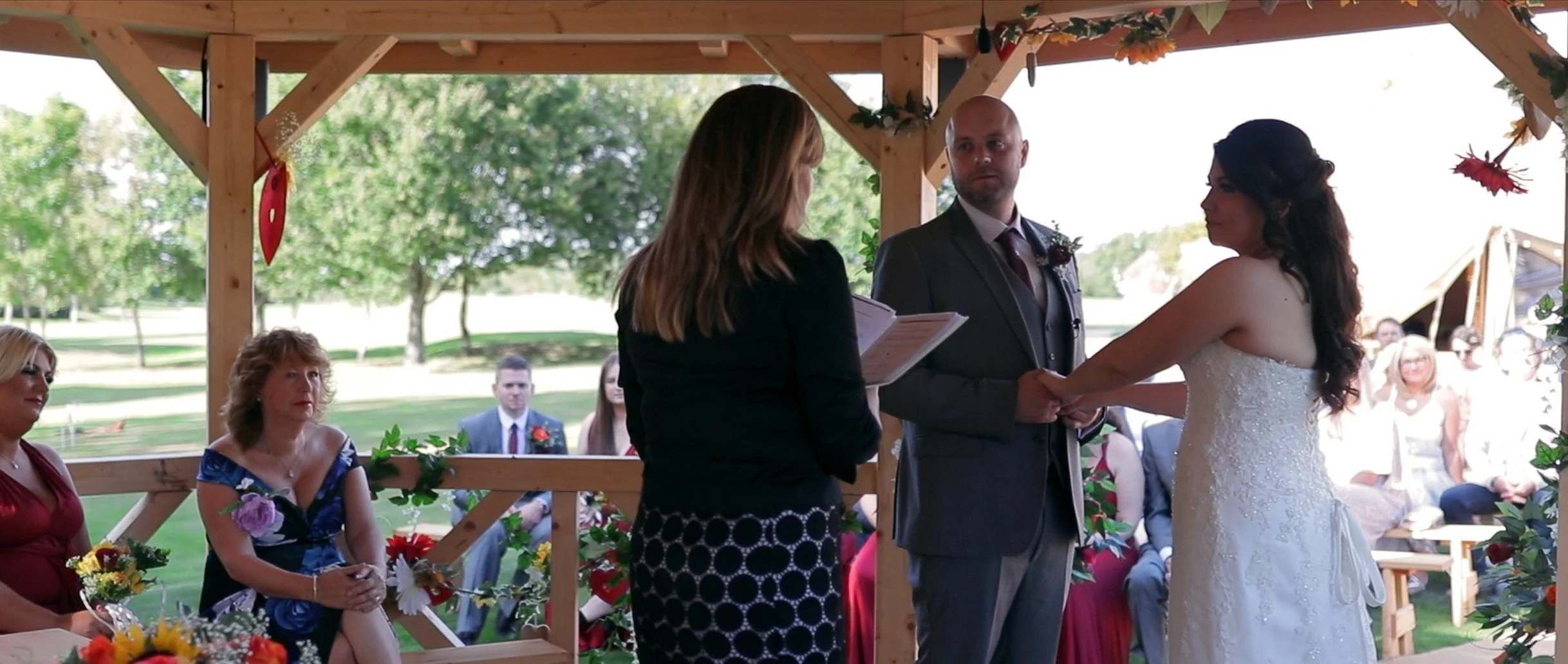 A wedding ceremony taking place outdoors under a wooden pavilion, with the bride and groom holding hands and facing each other, and an officiant reading from a book. Guests are seated in the background, with some women wearing colorful dresses and me
