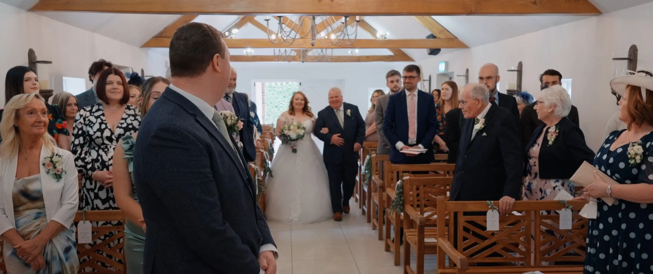 Wedding ceremony inside a small chapel with guests standing and facing the bride and groom. The bride is walking down the aisle towards the groom. The ceremony is decorated with flowers on the pews, and the ceiling has wooden beams and a chandelier.