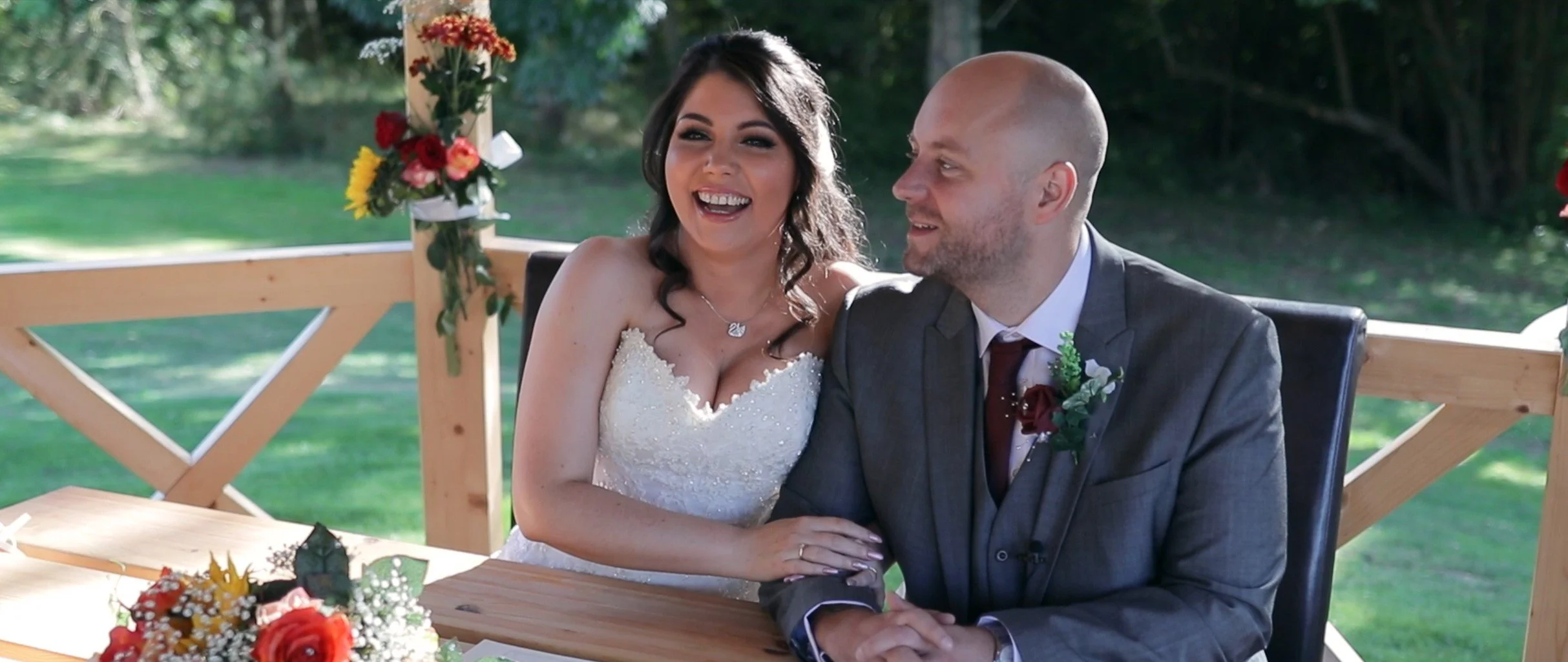 Bride and groom sitting at a wedding reception table outdoors, smiling and holding hands, with flowers on the table and a wooden railing behind them.