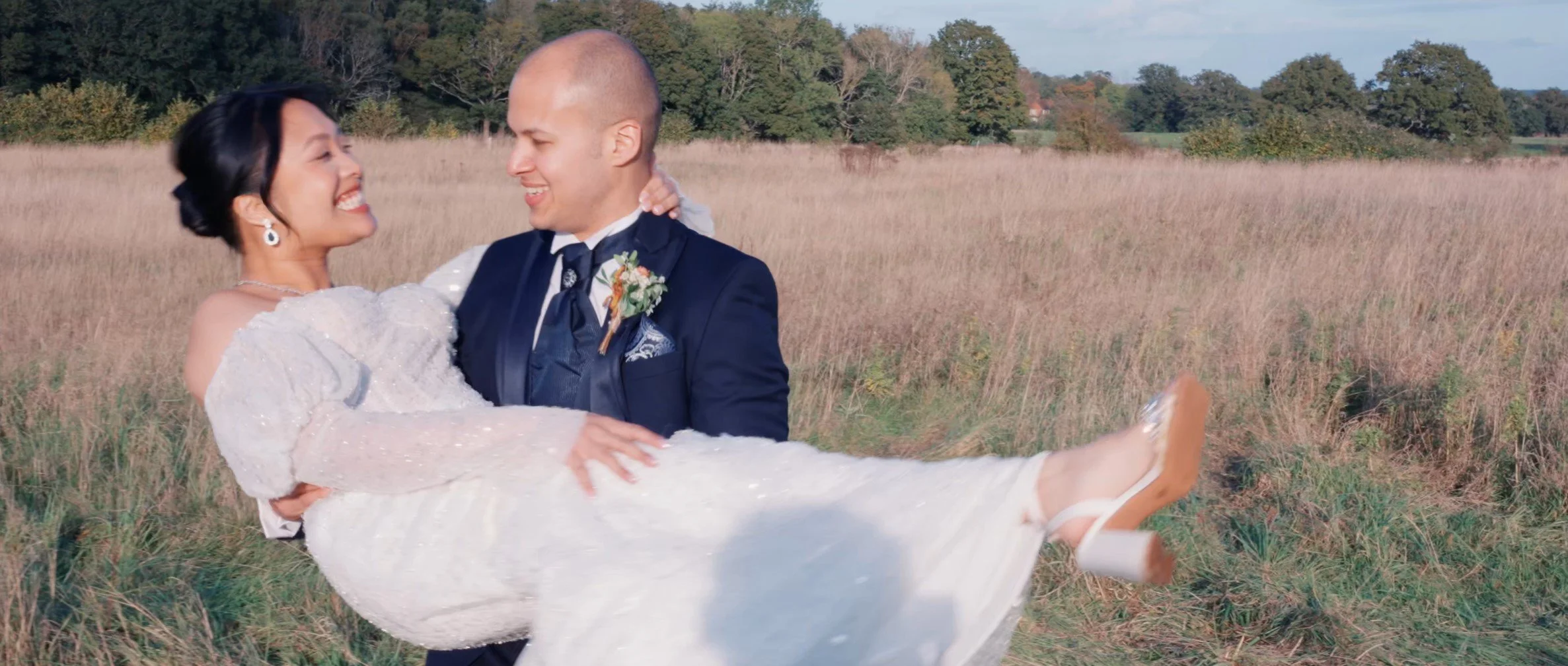 A newlywed couple in wedding attire outdoors, with the groom holding the bride in his arms in a field with grass and trees in the background.