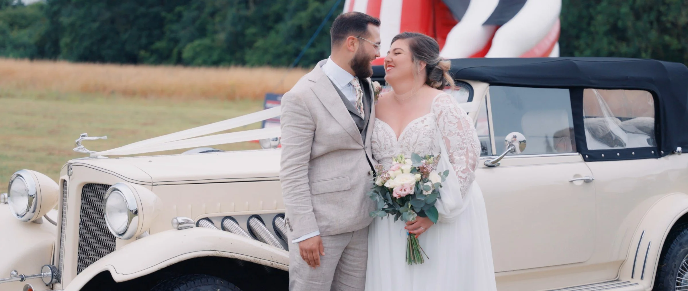 A bride and groom kissing in front of a vintage white car decorated with white ribbons, on a grassy field with trees in the background.