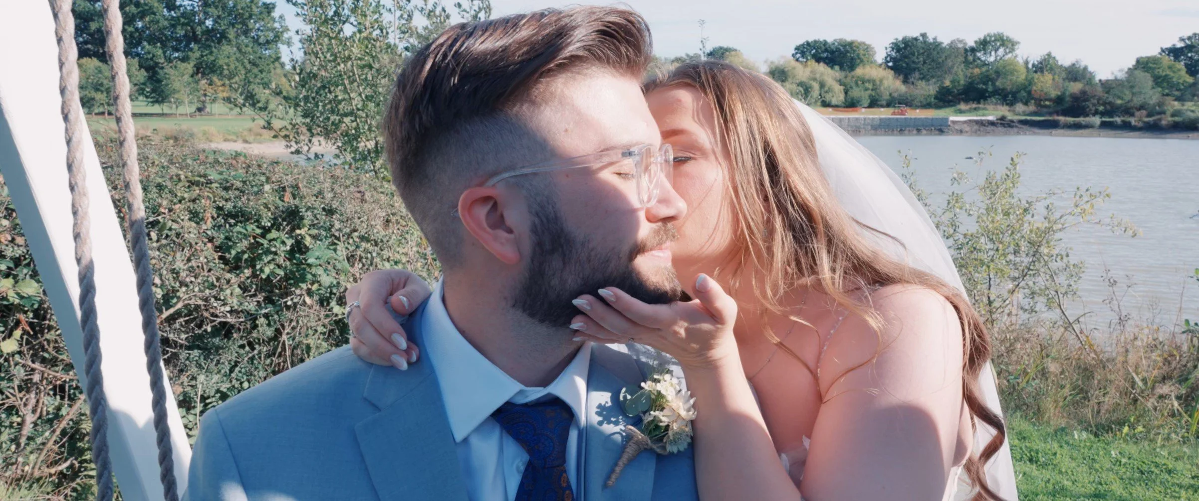 A couple sharing a kiss outdoors near a lake, with the woman gently holding the man's chin, both dressed in wedding attire, during daytime.