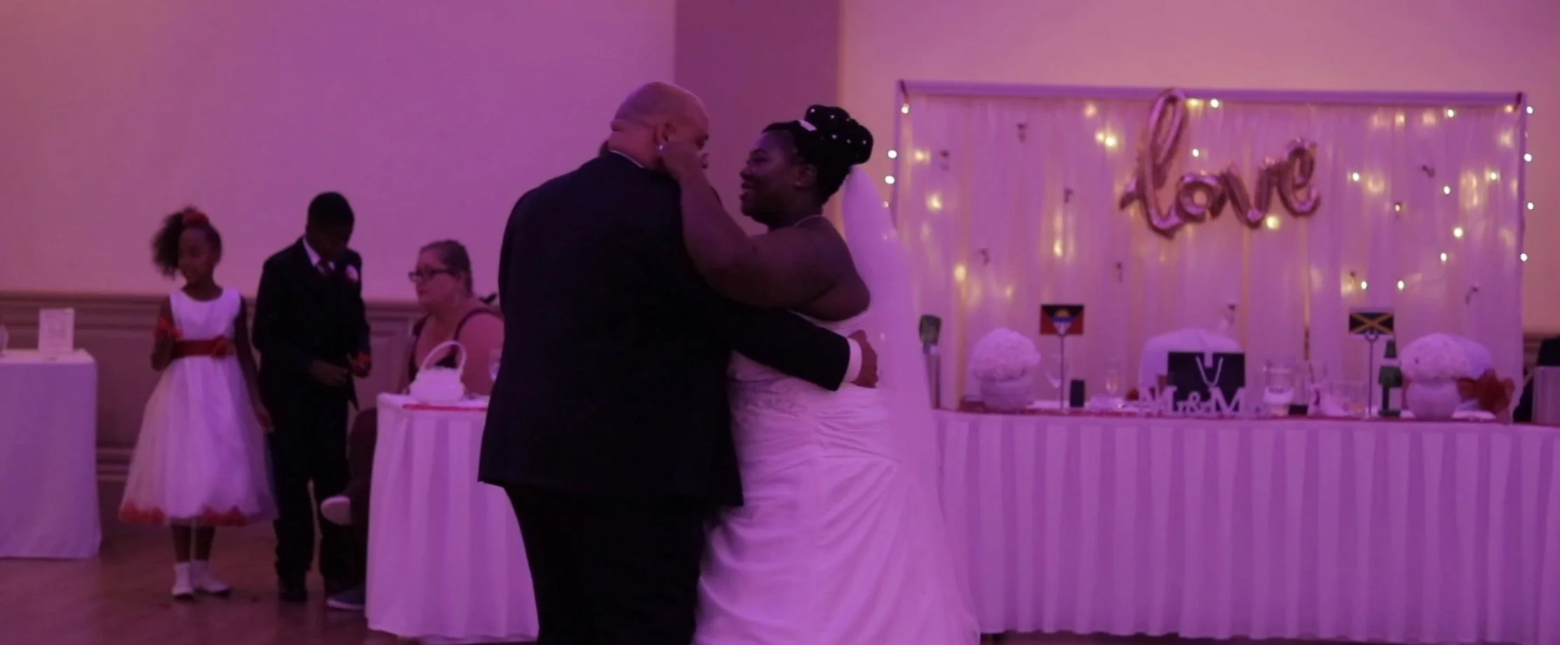 A couple dancing at a wedding reception with a decorated backdrop that says 'love' and has fairy lights, with some children standing nearby and a table with gifts and decorations in the background.