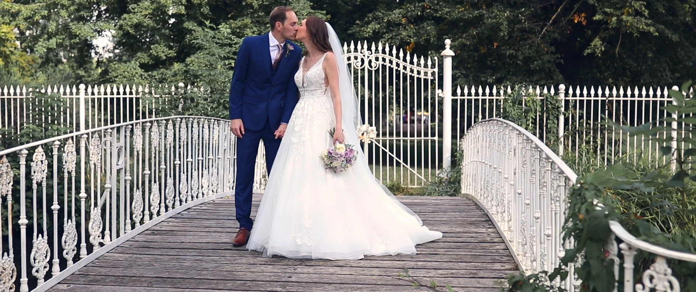 A bride and groom share a kiss on a small wooden bridge surrounded by greenery and an ornate white fence, with the bride holding a bouquet of flowers.