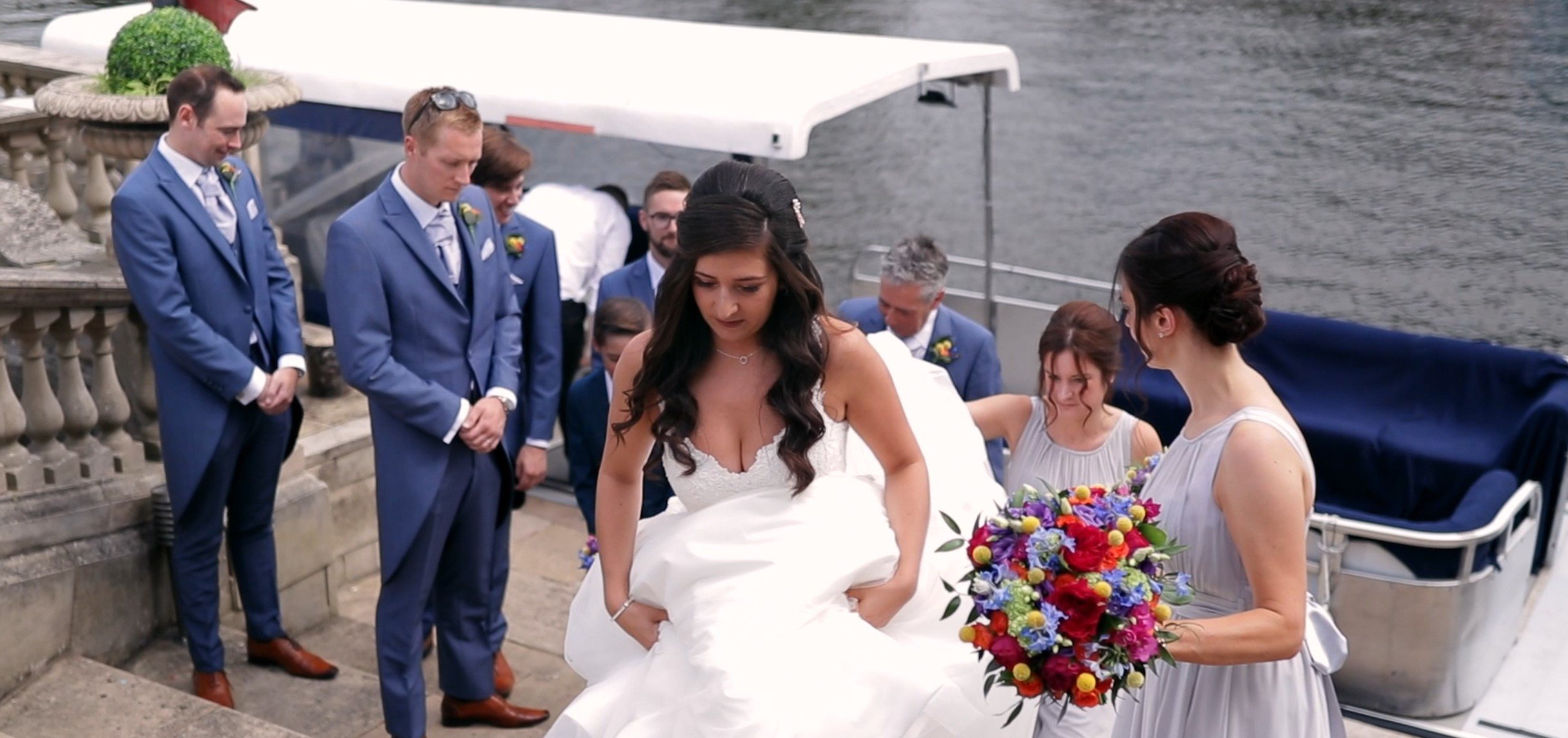 A bride in a white wedding gown and dark hair with loose curls, a bridesmaid in a light gray dress holding a colorful bouquet, and several groomsmen in matching blue suits with striped ties, standing near a boat on the water for a wedding ceremony.