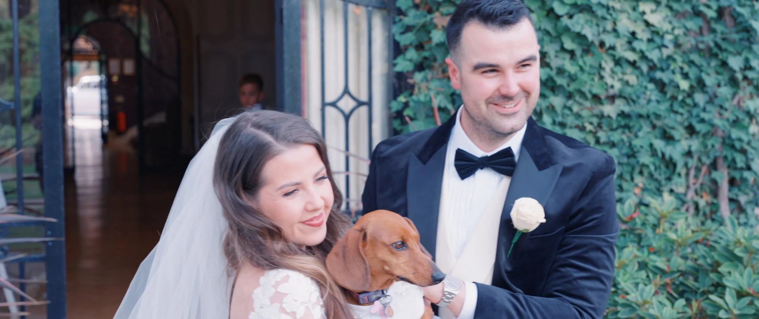 A bride and groom smiling with a brown dog at their wedding outside, with greenery behind them.