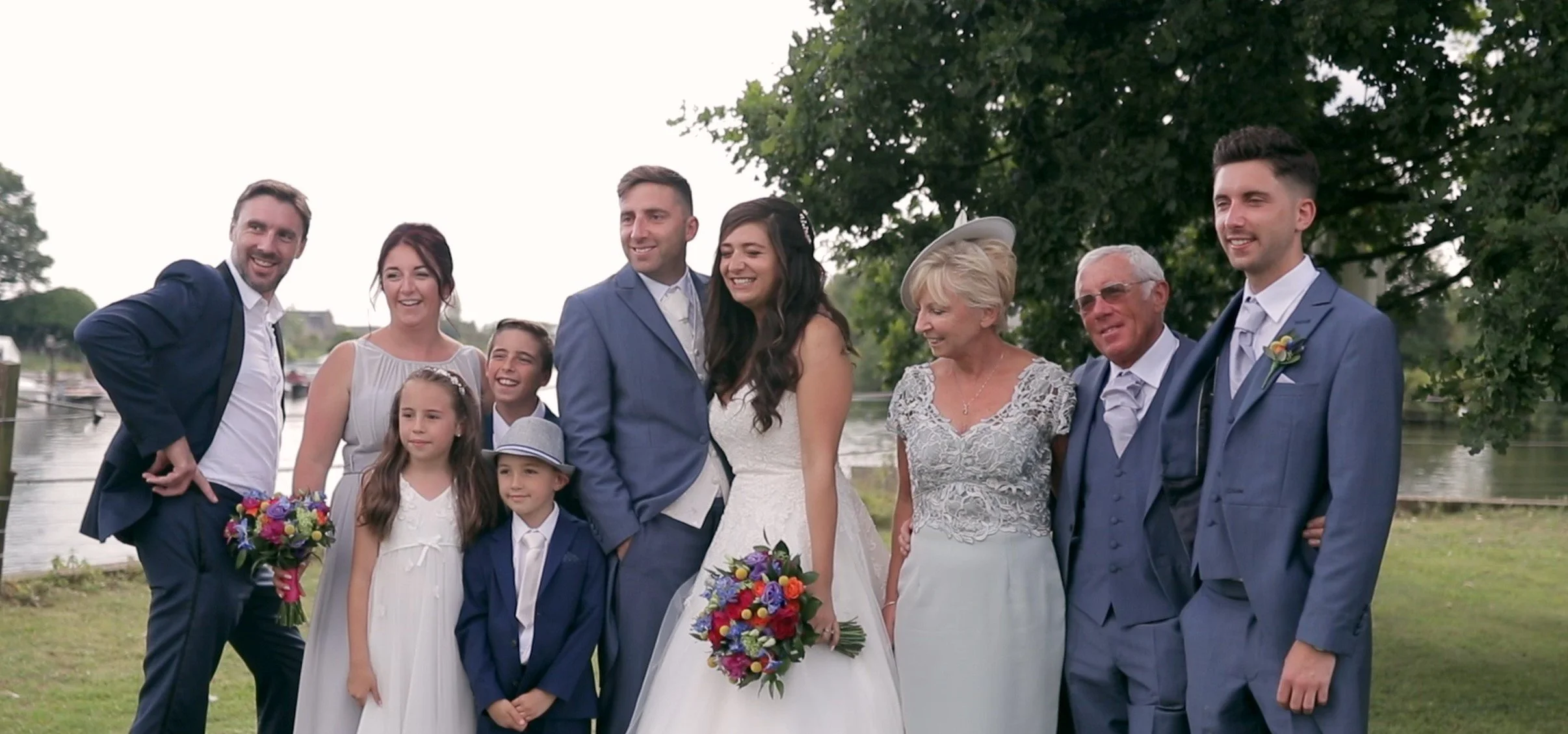 Group of people at a wedding by a lake, including a bride and groom in the center, surrounded by family and friends, all smiling and dressed in formal attire.