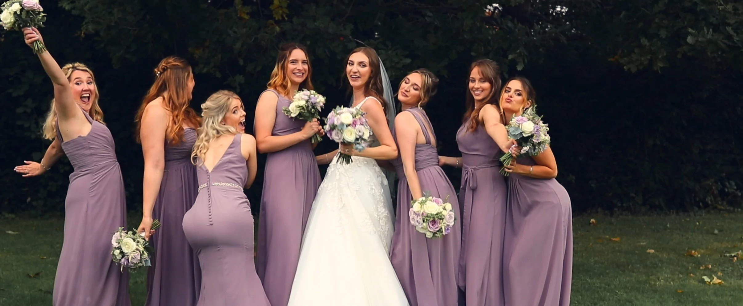 Group of women at a wedding, with two women in wedding dresses and six bridesmaids in lavender dresses, holding bouquets, posing outdoors.