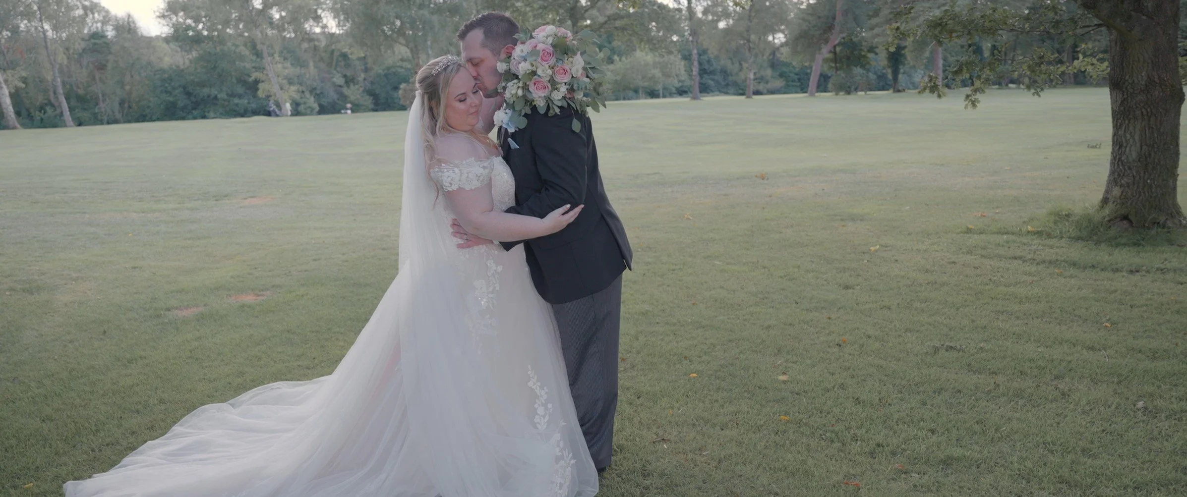 A bride and groom embrace in a park, with the bride wearing a white wedding dress and veil, holding a bouquet of pink and white roses, and the groom in a dark suit. They are standing on grass near a tree, with trees and open space in the background.