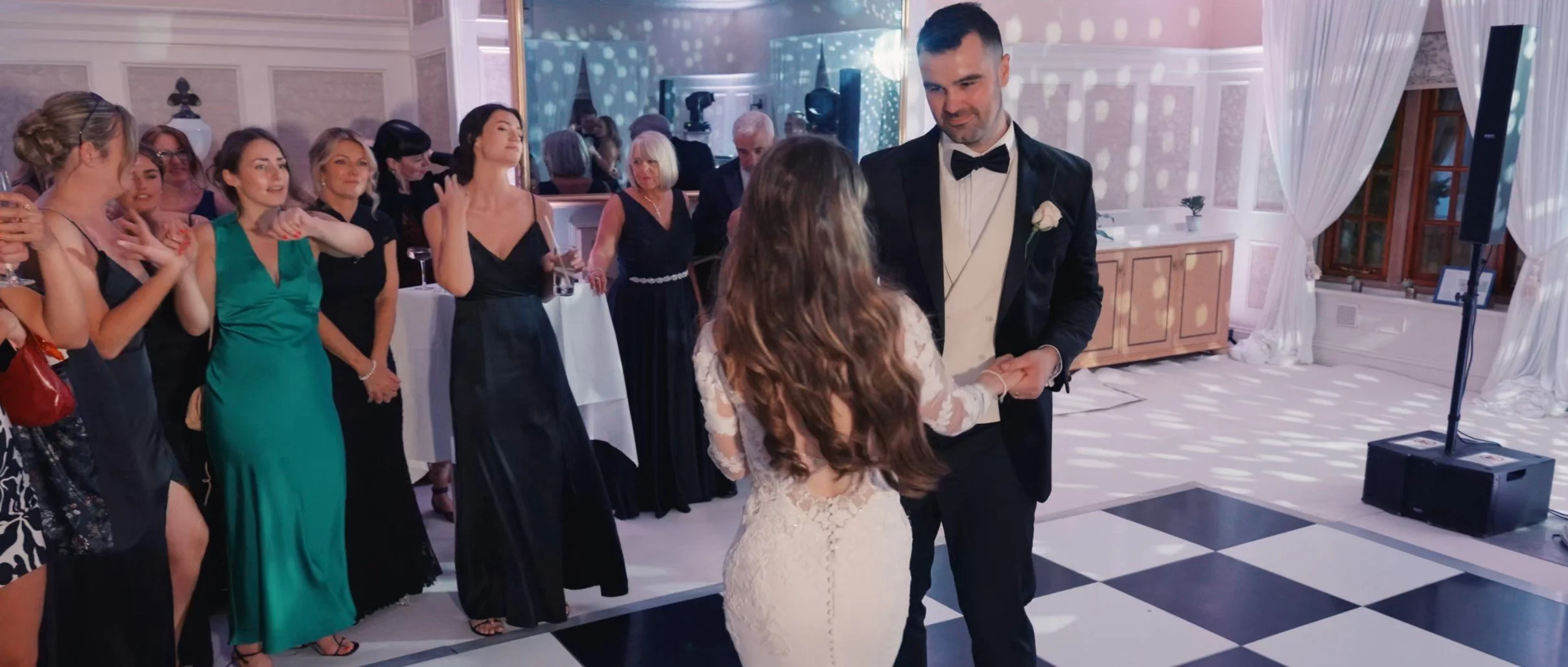 A wedding reception with a groom dancing with a young girl, possibly a flower girl or junior bride, while surrounded by female guests in formal dresses. The room has elegant decor with checkered black and white flooring, white drapes, and a large mir