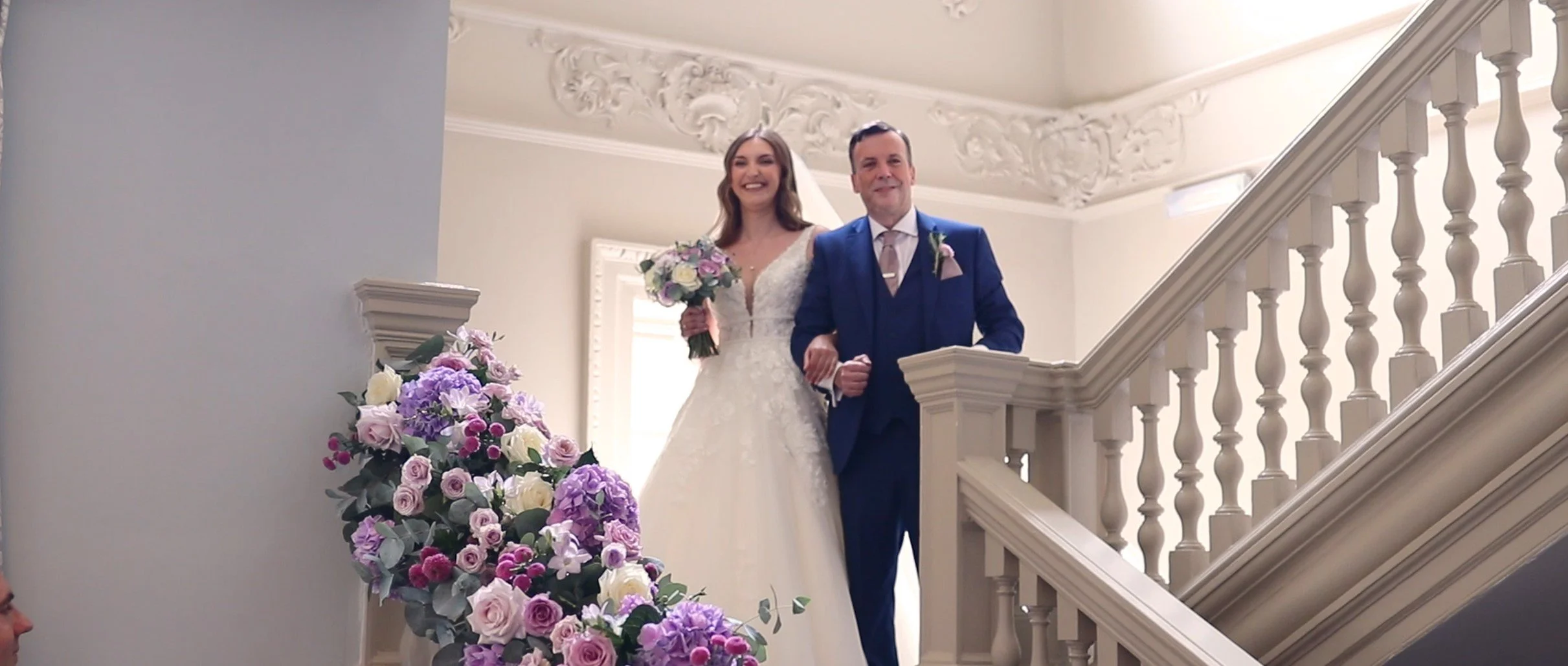 A bride in a white wedding gown and a man in a blue suit walking down a staircase, holding hands, with a bouquet of flowers in her hand. There are purple, pink, and white flowers on the side of the staircase, and ornate white moldings on the ceiling 