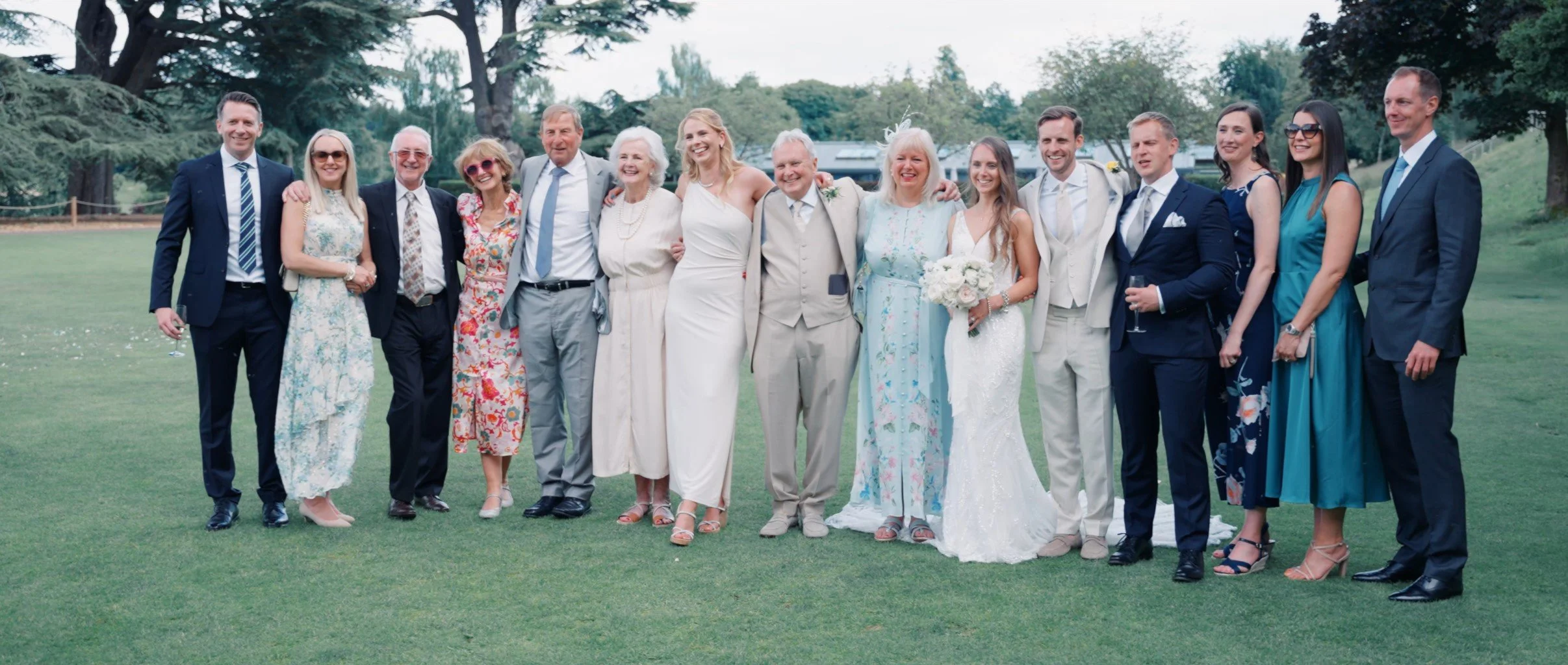 A group of 15 people, including a bride in a white wedding dress holding a bouquet, and others in formal attire, standing outdoors on a grassy area with trees in the background during a wedding celebration.