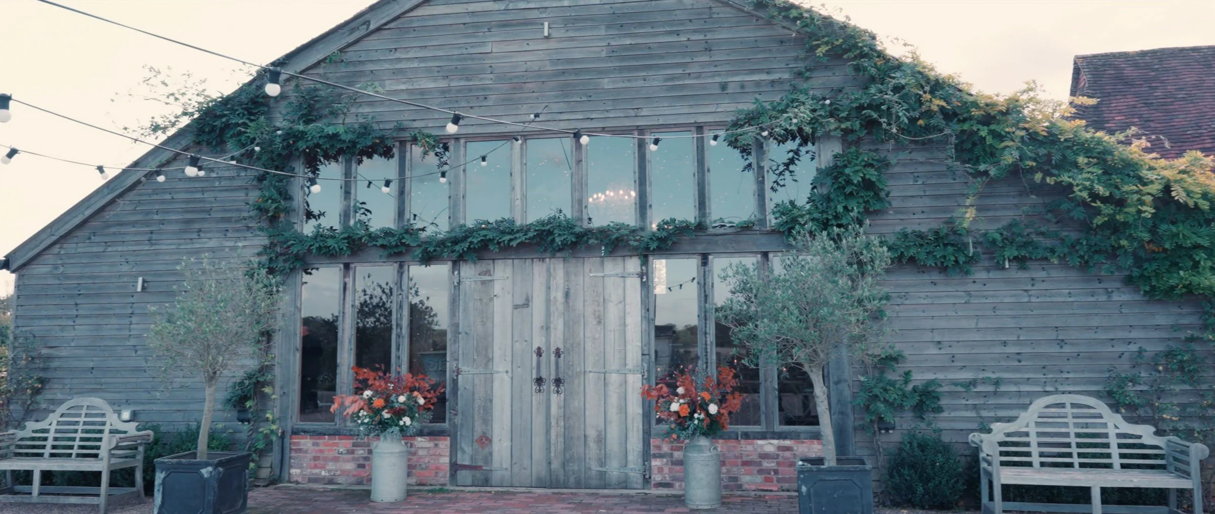 A rustic, two-story wooden barn with large front doors, surrounded by potted plants, flowers, and trees. String lights are hanging above, and there are two white benches on either side of the entrance.