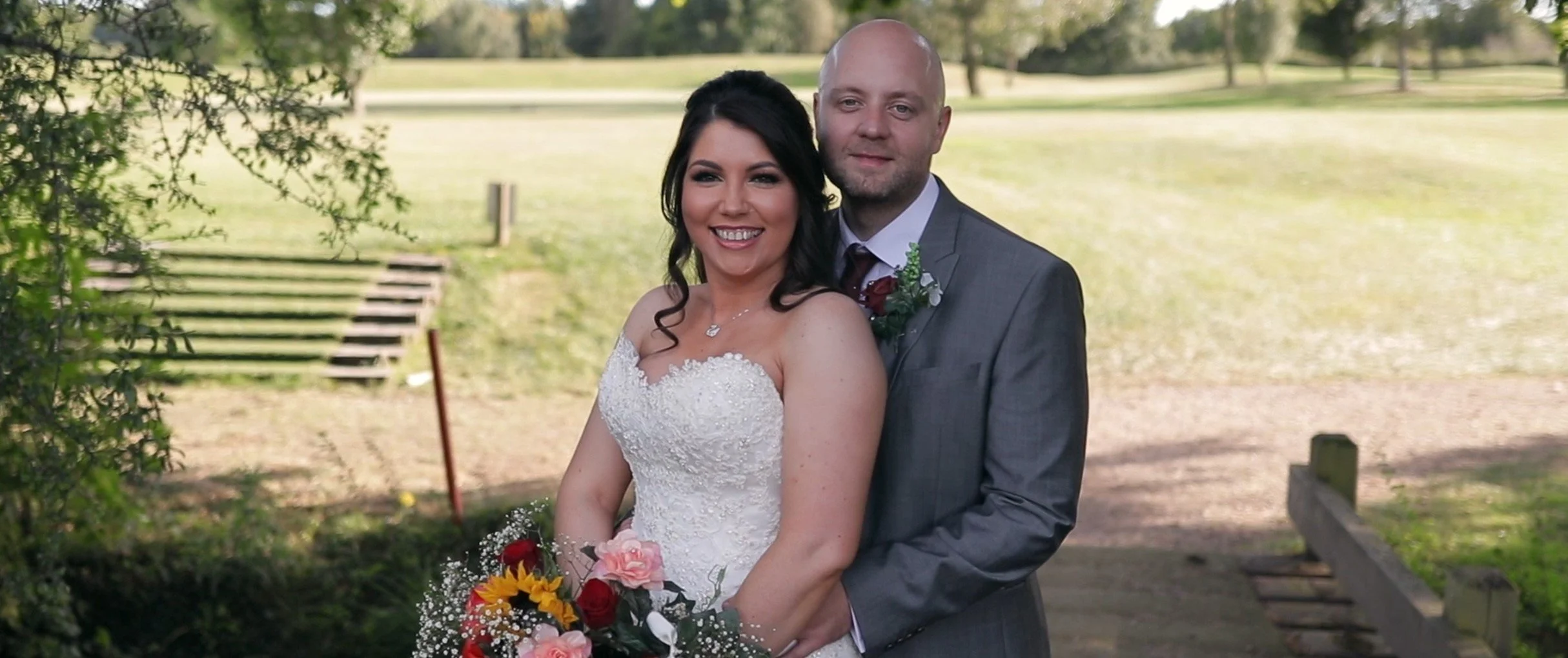 A bride and groom standing outdoors on a grassy area near stairs, smiling and posing for a wedding photo. The bride is wearing a white strapless wedding gown and holding a bouquet of flowers, while the groom is dressed in a gray suit with a white shi