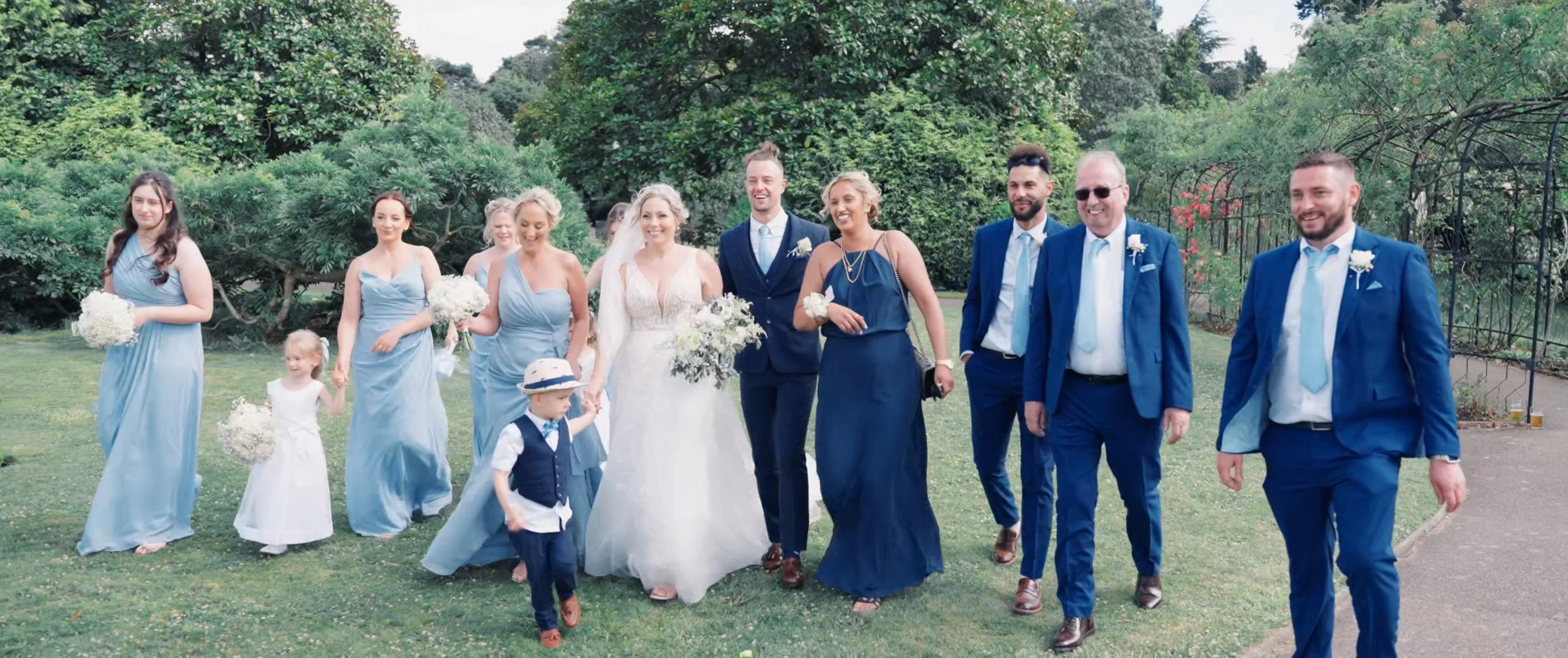 A wedding party walking outdoors on green grass with trees in the background, including bridesmaids in light blue dresses, a bride in a white wedding dress, groomsmen in blue suits, and children in formal attire.
