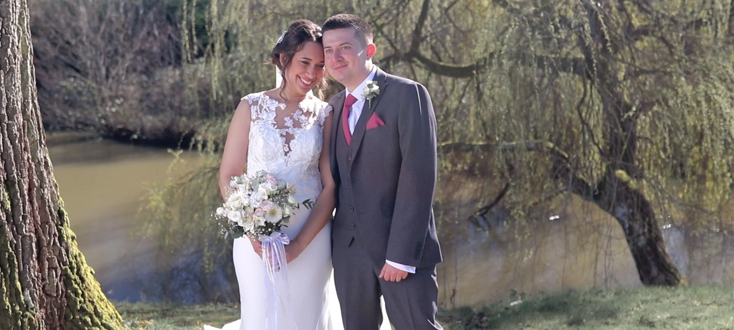 A bride and groom standing close together outdoors, smiling. The bride is holding a bouquet of white and pink flowers, and the groom is wearing a dark suit with a pink tie and boutonniere. Trees and a body of water are visible in the background on a 