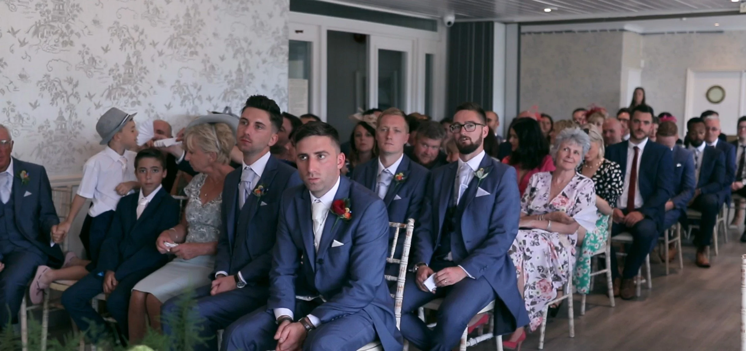 Wedding guests sitting in a decorated indoor venue, some in suits and formal dresses, watching the ceremony attentively.