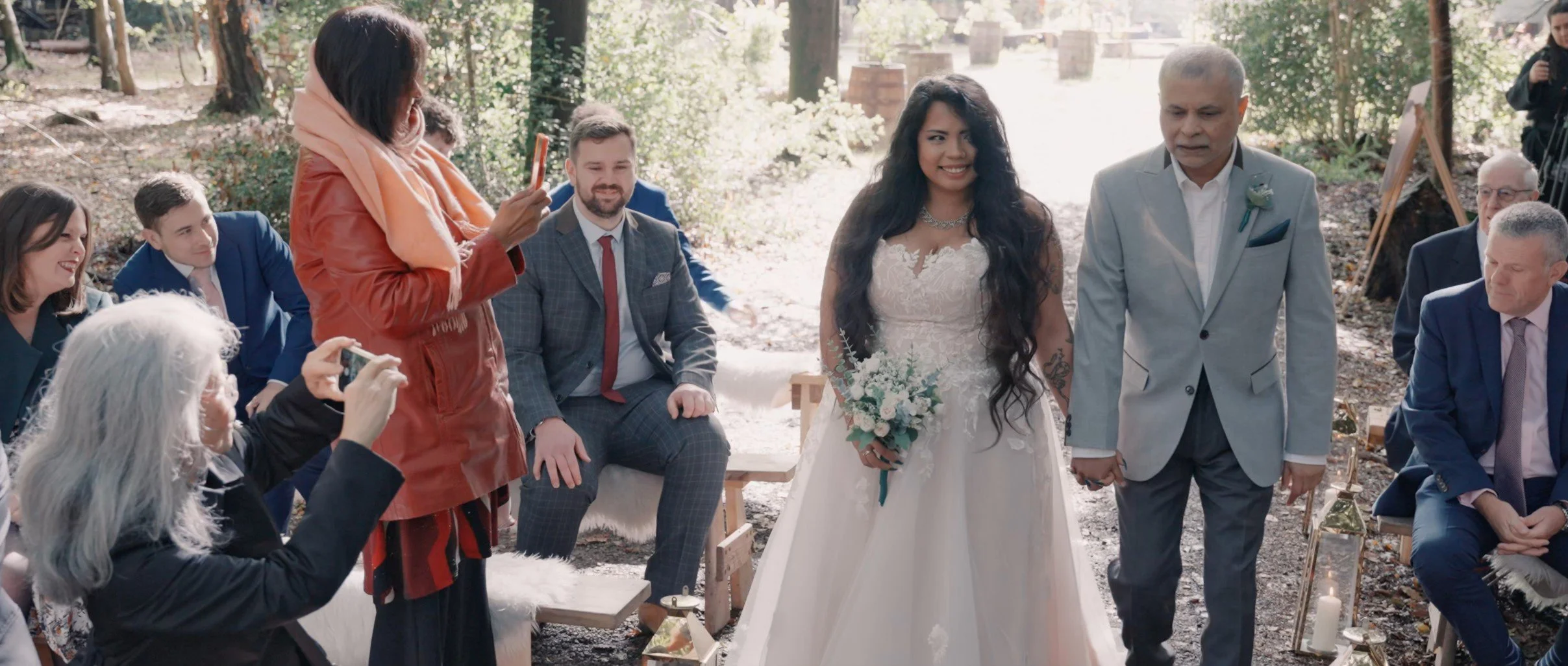 A bride in a white wedding dress holding a bouquet, walking down the aisle with a man in a light gray suit, during an outdoor wedding ceremony in a wooded area. Guests seated on either side are taking photos and watching the couple.