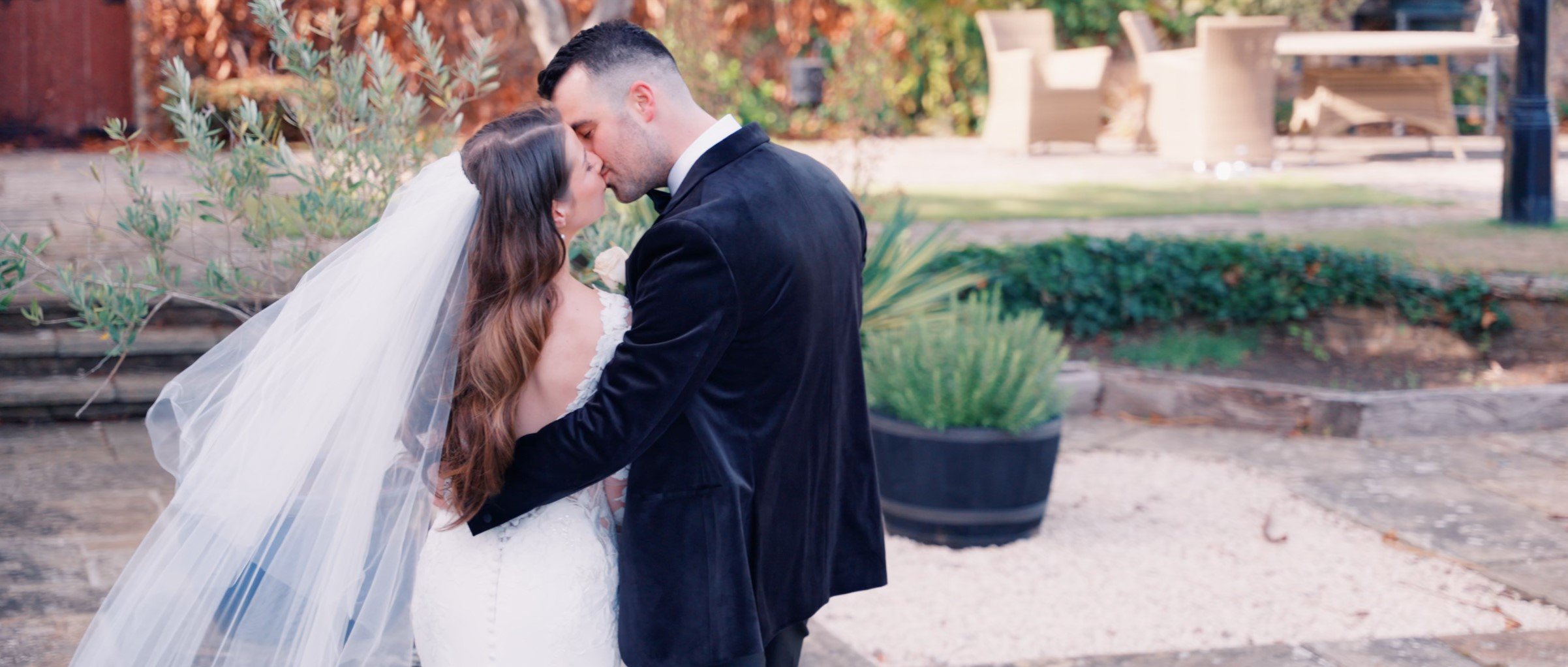 A newlywed couple sharing a kiss outdoors during their wedding, with the bride wearing a white wedding dress and veil, and the groom in a black tuxedo, surrounded by gardening plants and park benches.