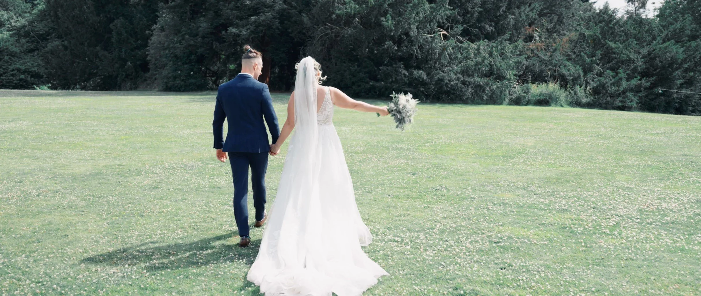 A bride and groom walking hand in hand across a grassy field during their outdoor wedding, with the bride holding a bouquet and wearing a white dress, and the groom in a dark suit. Trees are visible in the background.