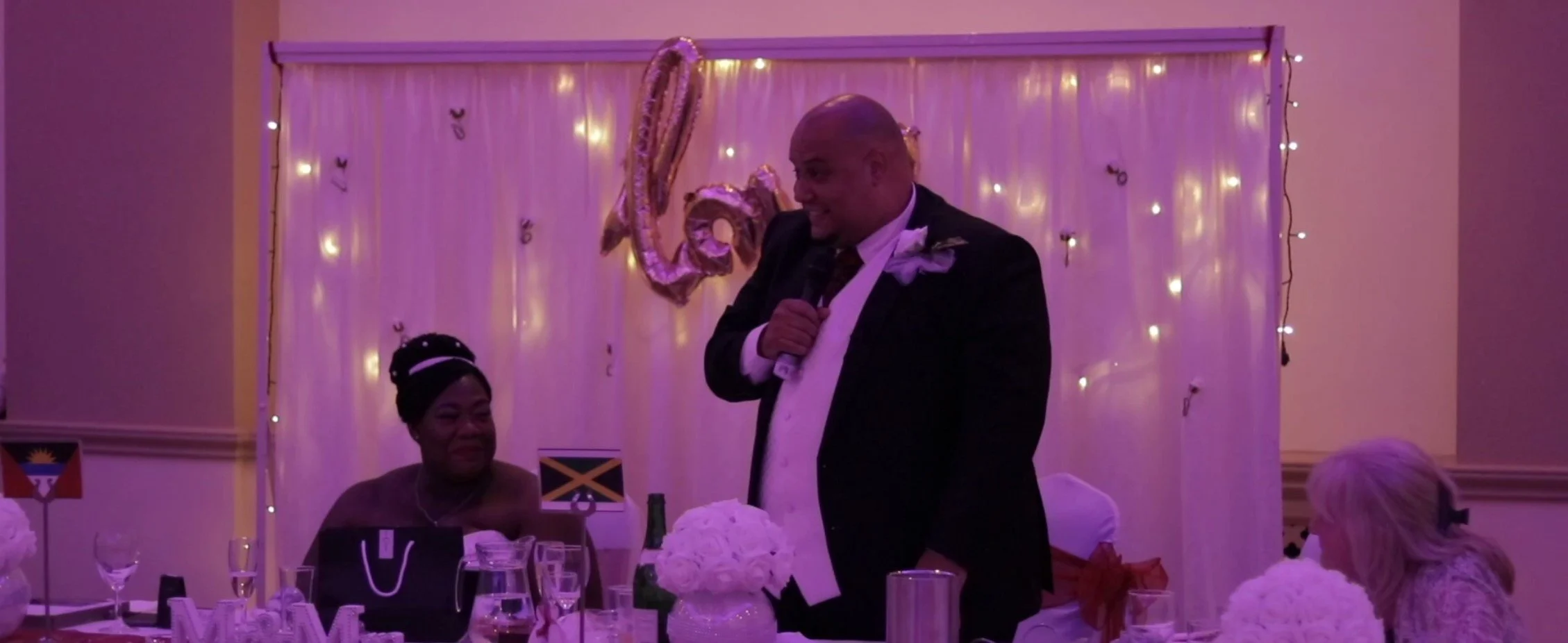 Man in black tuxedo giving a speech at a wedding reception with a microphone, surrounded by seated guests, floral arrangements, and purple and gold decorations.