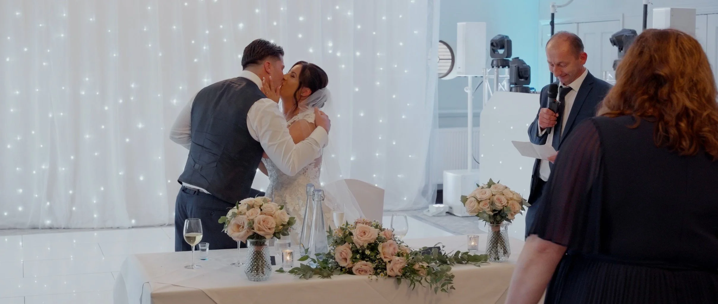 A bride and groom share a kiss at their wedding reception, with an officiant reading and floral decorations on the table.
