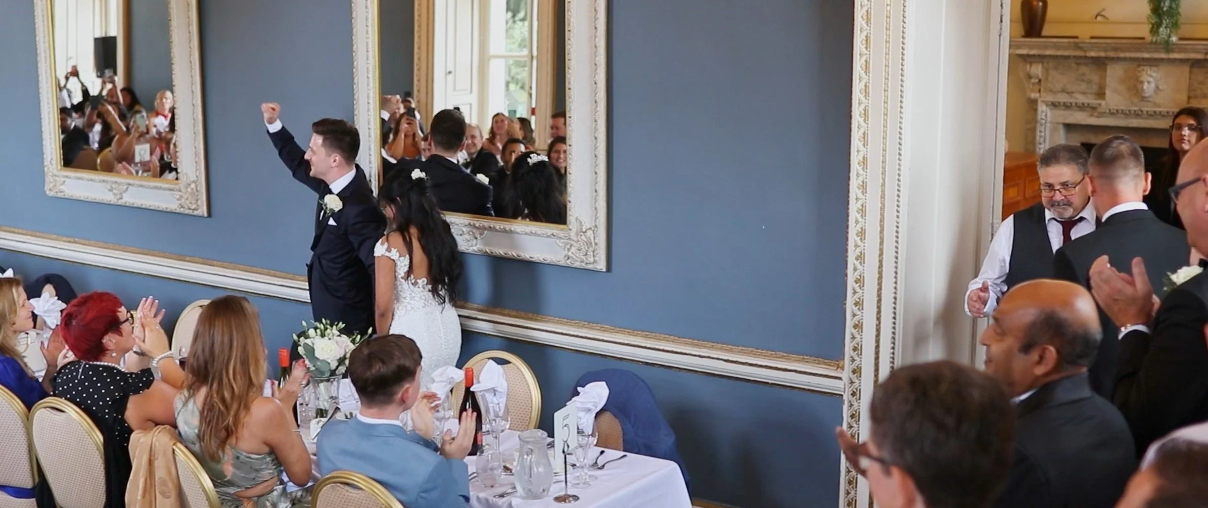 A wedding reception with guests seated at tables and a bride and groom standing, surrounded by family and friends, with some clapping and others taking photos.