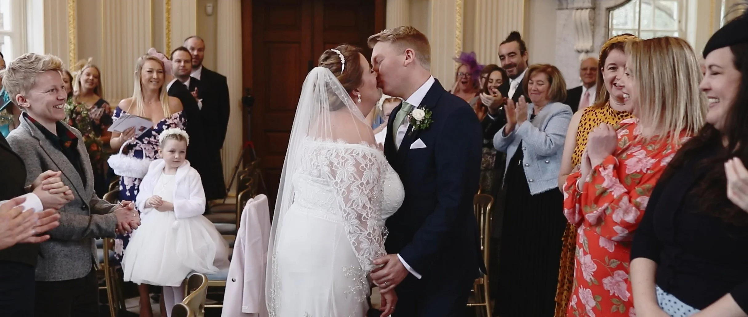 A bride and groom kiss during their wedding ceremony, surrounded by smiling guests clapping and celebrating indoors in a decorated hall.