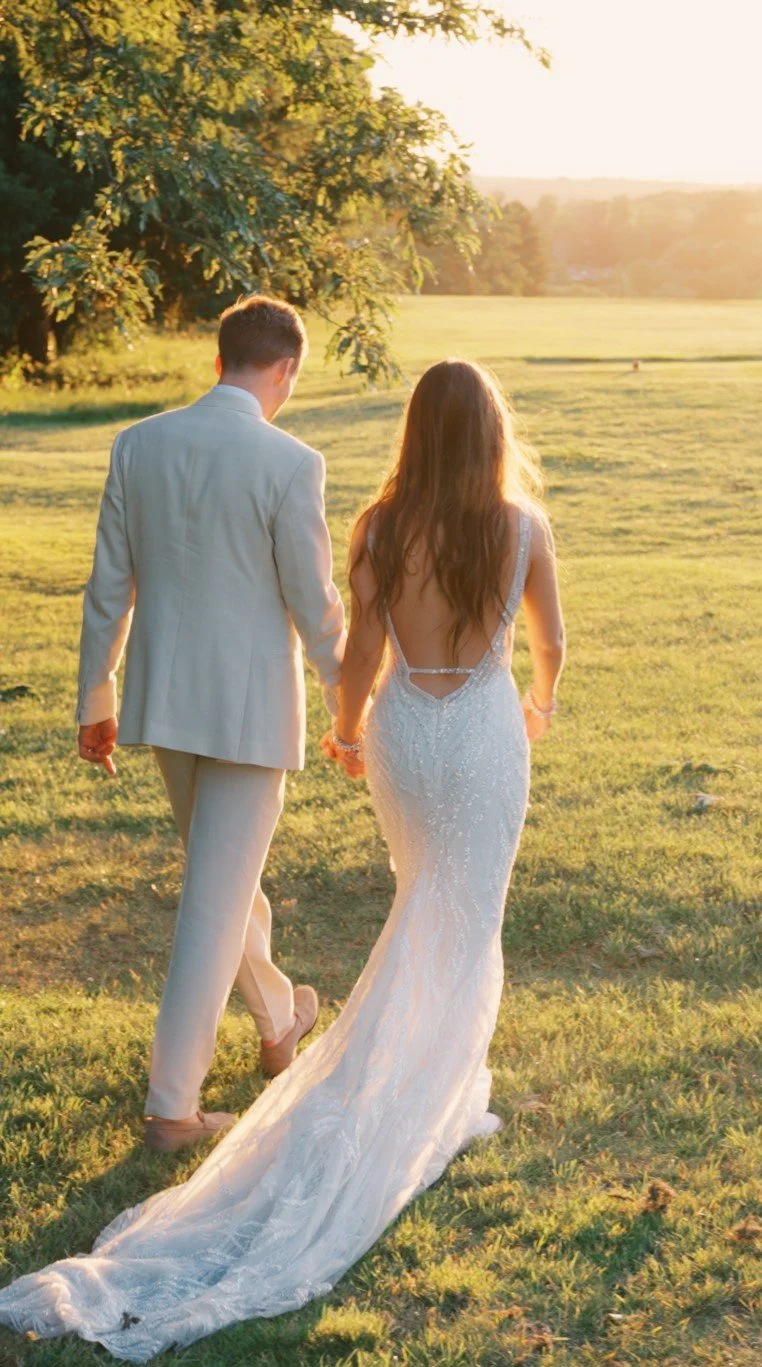 Bride and groom walking through open fields at sunset on their wedding day, captured in a natural style.